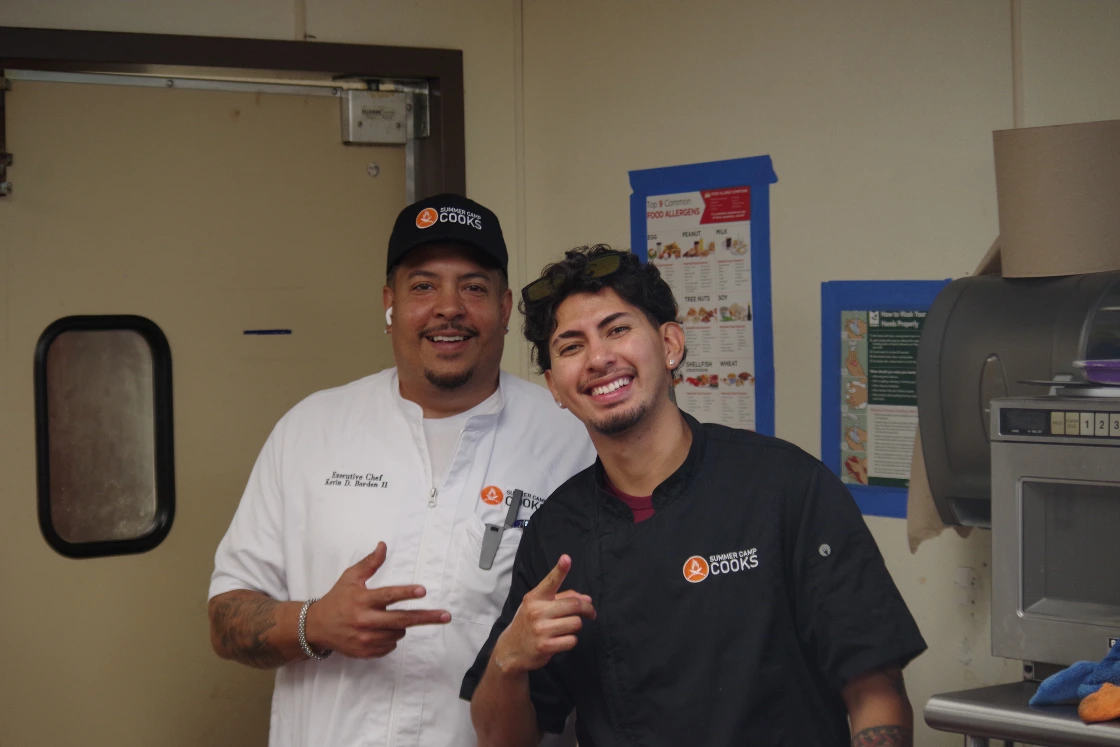 Two Summer Camp Cooks chefs smiling in a camp kitchen, with a food allergen chart posted on the wall behind them