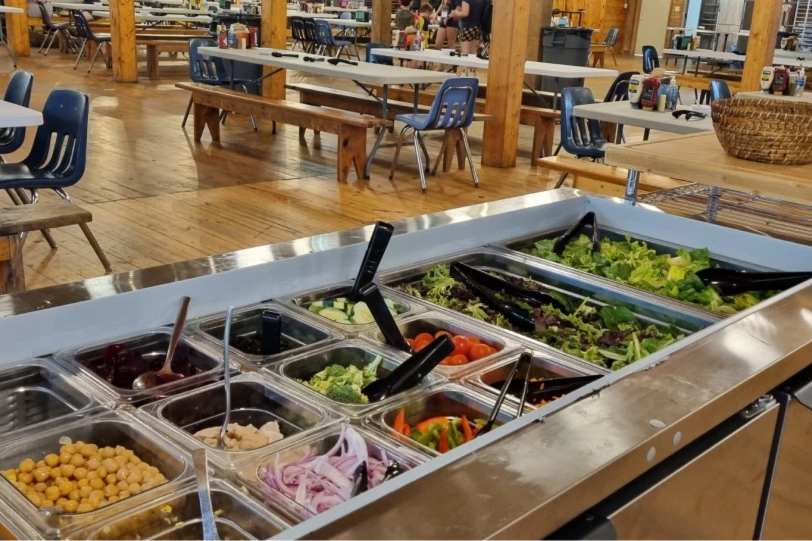 A camp dining hall serving line stocked with fresh fruit, vegetables and salad components